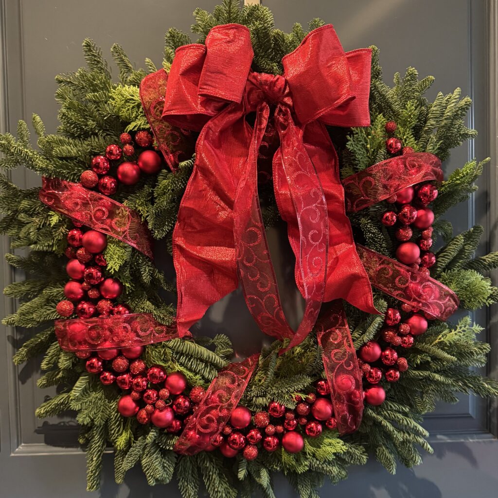 Handcrafted Classic Noël holiday wreath featuring fresh evergreens, elegant red ornaments, and a luxurious satin ribbon.